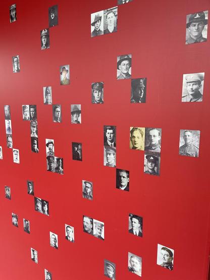 This image shows a red wall that is decorated with numerous black and white photographs of individuals. These photographs vary in size and seem to be portraits of men, possibly from different time periods, as indicated by their attire and the styles of the photographs. Some of them are wearing military uniforms. The display could be part of a historical exhibit, memorial, or a thematic art installation aiming to convey a specific message or evoke a certain era.