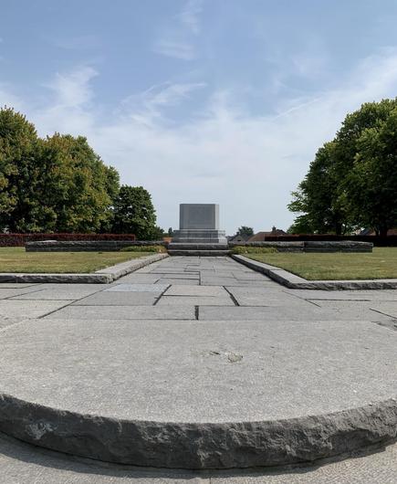 The image shows a symmetrically aligned pathway leading up to a monument or memorial. The monument is centered at the far end of the pathway and appears to be a large rectangular structure with inscriptions on its front. The setting is peaceful, surrounded by well-kept green lawns and mature trees. The sky is blue with some scattered clouds. Based on the structure and surroundings, this location may serve as a dedicated space for remembrance or tribute.