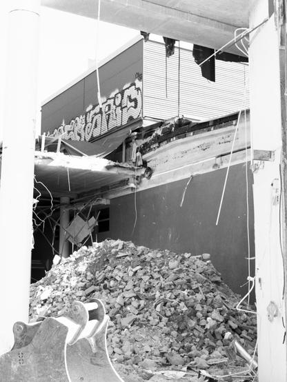 Black and white image of a building under demolition, with a pile of rubble in the foreground. The surroundings are partially collapsed, exposing tangled wires and graffiti on a wall.