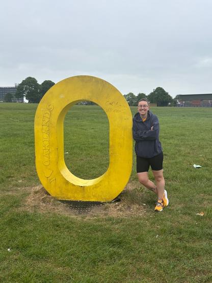 Photo of Emily standing next to a large (taller than her) 3D O. It is in a large grass area. She is nonchalantly leaning on the O like she is in a photo shoot for a catalogue.
