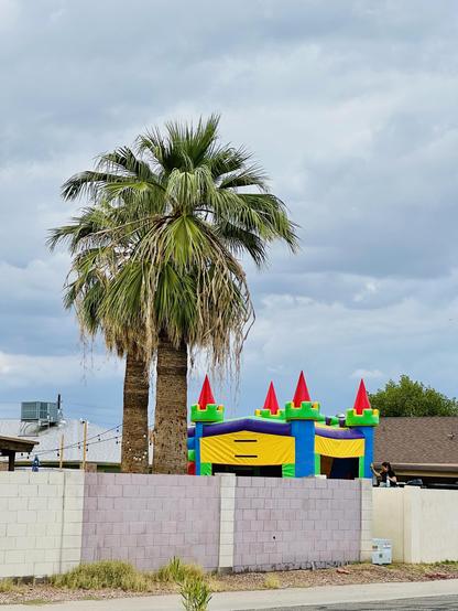 Seen over a neighborhood cement block wall, a giant Bounce House castle rising up about 16’ tall and maybe 12’ wide, primary colors. Under 2 palm trees with full canopy because they haven’t been trimmed in years. The blue gray sky is filled with clouds so the bright children’s party castle stands out.