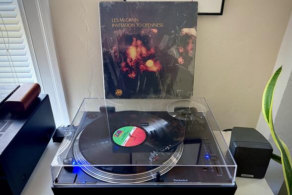 A black vinyl record spins on a black turntable. The record sleeve stands up on the turntable’s closed dust cover behind the platter.