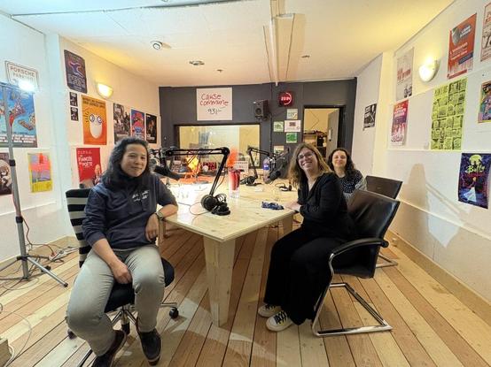 Florence, Emmanuelle et Céline (de gauche à droite sur la photo) autour de la table en bois du studio. Sur la table on voit 4 micros. Il y a des affiches sur les murs du studio.