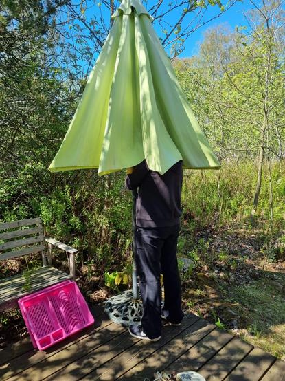 Allekirjoittanut seisoo mökin terassilla ison vaaleanvihreän aurinkovarjon sisällä. Vieressä jätelavalta pelastettu kirkkaan pinkki astiankuivausteline.