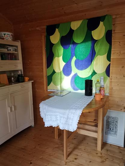 Kuva mökin sisältä. Ikkunalla on retro Marimekon vihreän sävyinen Villikaali-verho, klaffipöydällä on pitsiliina ja ruokatarpeita, ja vasemmalla näkyy emännänkaappi, jossa kaikenlaista keräilytavaraa.