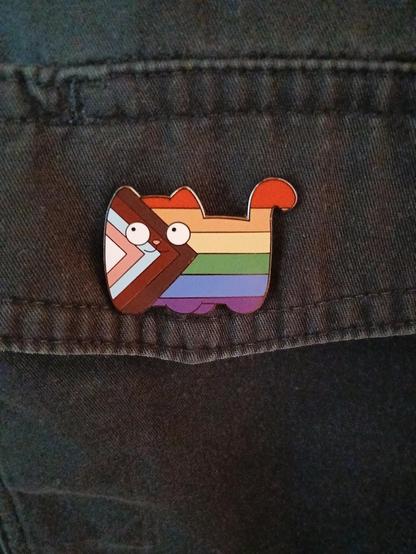 Pin in the form of a cat in the inclusive pride flag colors on a black shirt. The cat has cute googly eyes.