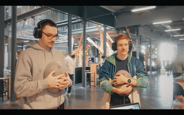 L'image représente deux personnes (hommes) qui testent chacun une enceinte Pacini 8 en la serrant dans leurs bras et contre leur poitrine. Ils sont équipés d'un casque antibruit.