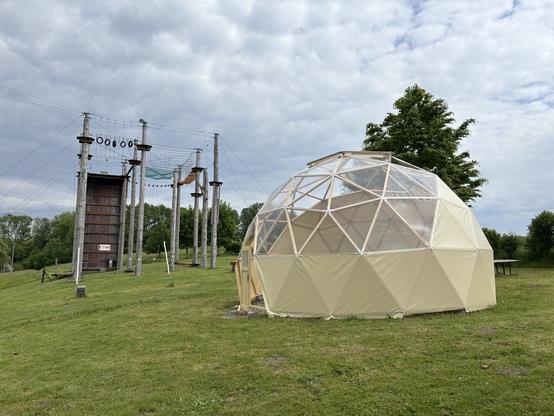 Ein Geodome - eine Halbkugel aus hölzernen Dreiecken mit Folie bespannt - vor einem Hochseilgarten. Auf einer grünen Wiese, der Himmel ist bewölkt.