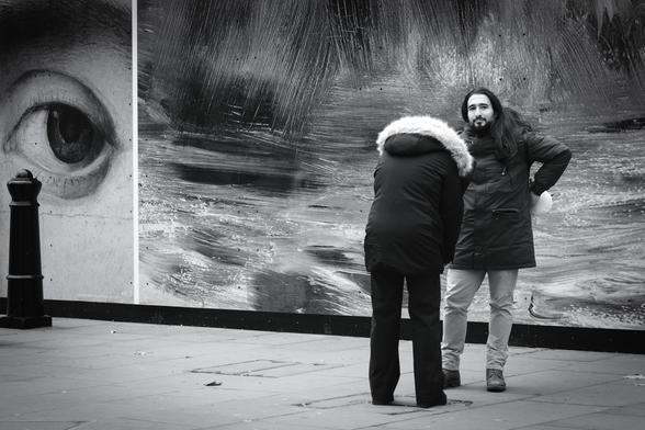 Black and white. A young couple are standing near very large photos of details from famous works of art. He is looking directly at my camera and not at the woman's phone as she takes a photo of him. At the far left of the picture, a large painted eye also seems to be looking at my camera.