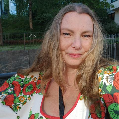 Head and shoulders selfie of a white middle aged woman wearing an off-white dress with 1970s flower pattern in green and warm read. She is smiling and has reddish light brown long hair with streaks of silver.