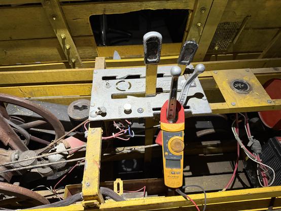 A clamp multimeter clamped over one of the lever controls in the middle of the vehicle, with the engine to the left and a red fuel tank to the right