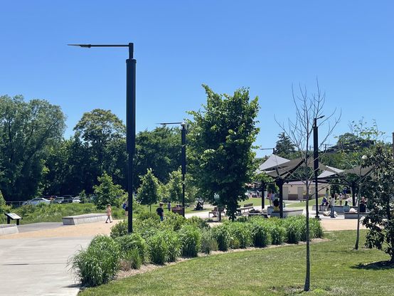Several light posts branch out beside a path winding through a town park. Solar panels sit on top on the light. Bushes and trees and grass sit below a clear blue sky.
