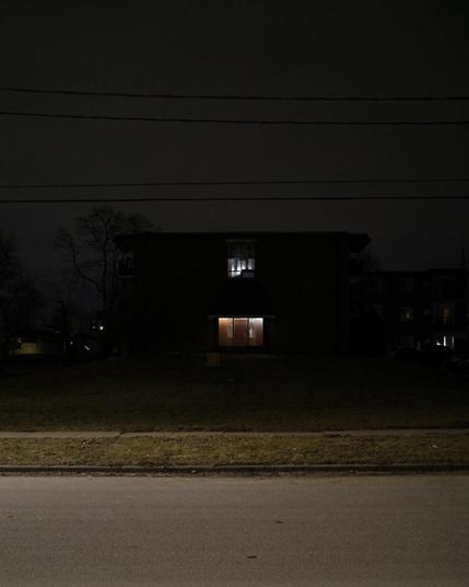 Color photograph looking across a well-lit empty street at night and across a lawn that recedes into shadow and darkness at a side entrance face of an apartment building, a dark blocky silhouette against a dark slate gray sky with lights shining from inside through a glass entrance and a window on the floor above it