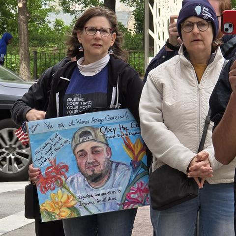 At a protest a woman holds a sign with a colorful drawing of Kilmar Abrego Garcia. It says Injustice for one of us is injustice for all of us.