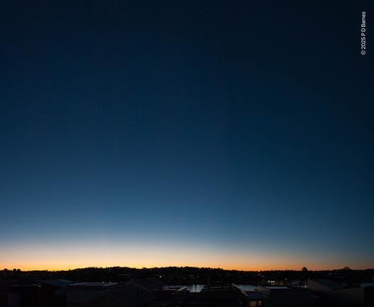 A perfectly clear, cloudless dawn sky. A dimly visible urban landscape runs across the bottom of the picture: rooftops, a river, an undulating ridgeline forming the horizon. Above, the sky is just beginning to brighten, a narrow band of peach-white, then above an infinite vault of ever-deepening blue, serene and limitless.