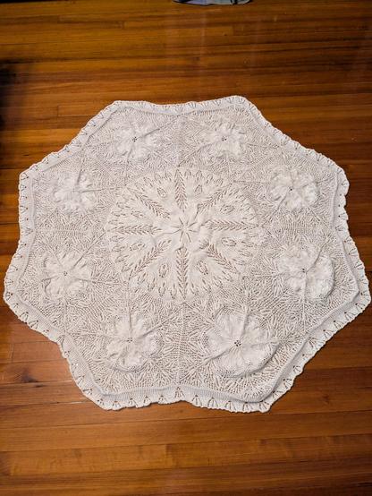 Large, white lace knit doily on a wooden floor. The doily is roughly octagonal, but it's rumpled and doesn't lie flat. Each corner has a large flower motif surrounded with festoons and an open mesh. The middle features a denser circle with 8 larger festoons radiating from the center.