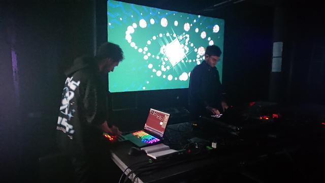 Two performers on laptops in front of a projection screen.