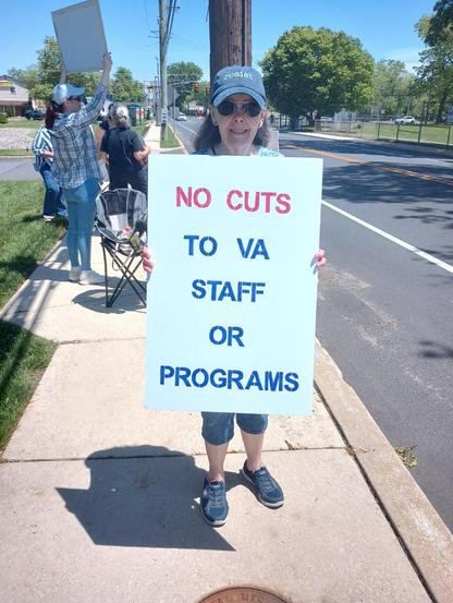 A woman with brown hair, and a hat that says, "Resist," is holding a sign on a sidewalk along a roadway.  The signs reads, "No cuts to VA staff or programs."