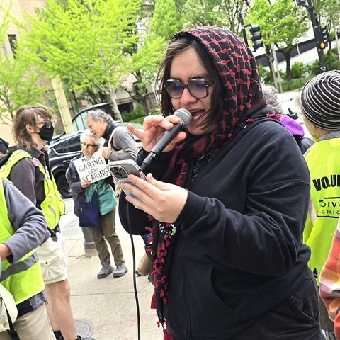 A woman wearing a scarf speaks into a microphone at an outdoor rally. In the background, another woman has a sign that says More caring, less scaring