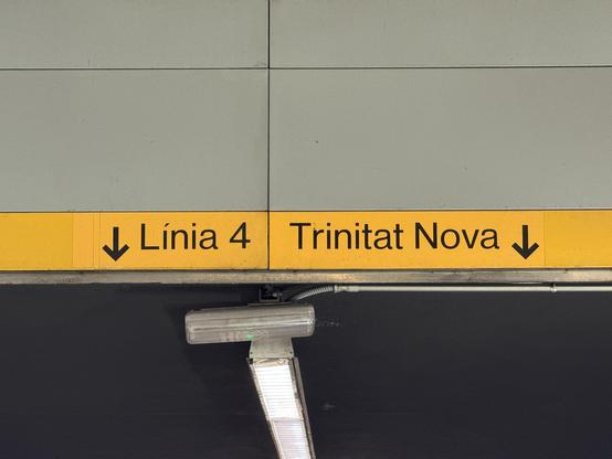 Wall with grey metal panels and a yellow metallic strip panes with a text in black between two arrows pointing down “Línia 4 Trinitat Nova” and below that it’s visible the ceiling with a strip of fluorescent lights and a emergency light.