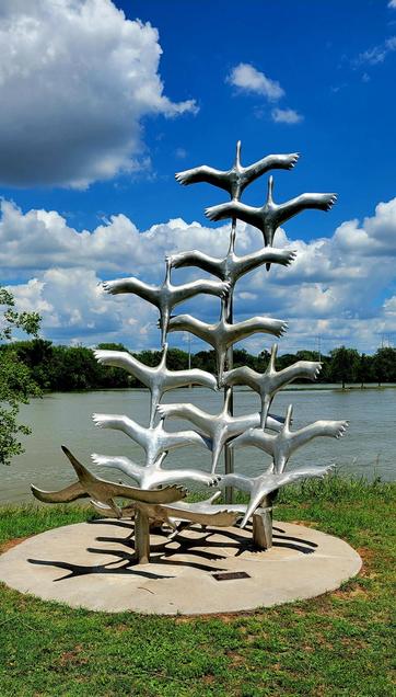 Chrome sculpture of a flock of geese taking flight. Installed on a concrete platform on river side. Blue skies with lots of white clouds above. Green tree line on the other side of the river.