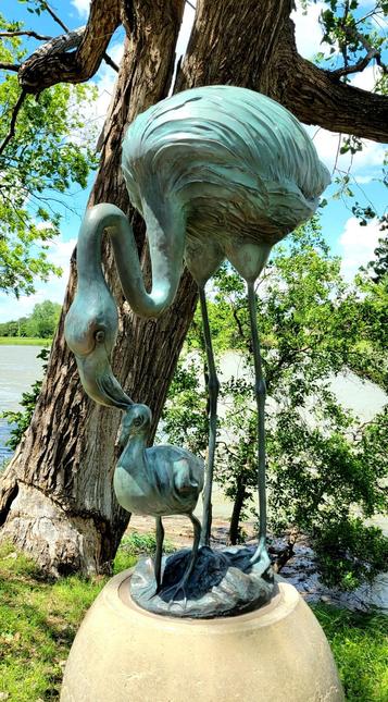 Green ("oxidized") sculpture of a mother flamingo bending down to pet its young one on the top of the head. Installation on barrel-shaped concrete platform on river side, next to a tree. Light blue sky with some white clouds visible behind the tree.