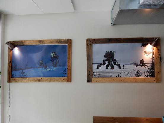 Two Simon Stålenhag art prints in wooden frames on a white wall:

Nattens Röntgenplåt (Two two-legged mechas walk through a dark blue night in a snowy landscape.)

Unlock. Explore. Repeat. (Two four legged, abanondoned military mechas get explored by two young people in a snowy landscape.)