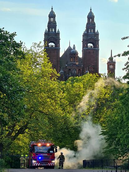 A fire engine in a Glasgow park with a firefighter outlined against smoke billowing from a large bin. Above it are the distinctive towers of the nearby Kelvingrove Art Gallery.