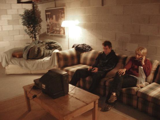 The image depicts a dimly lit living room with exposed concrete block walls. A plaid-patterned couch stretches across the right side of the room. Seated on the couch are two individuals, one holding a game controller and the other talking on a phone. In front of the couch, there is a wooden coffee table with an old-fashioned television set. On the left, a cushioned bench or small sofa is covered with a blanket, under which a figure is sleeping. A tall floor lamp beside the couch provides the main source of light, casting a warm glow over the scene. A large framed picture hangs on the far wall. A leafy plant is visible in one corner, adding a hint of greenery to the room.