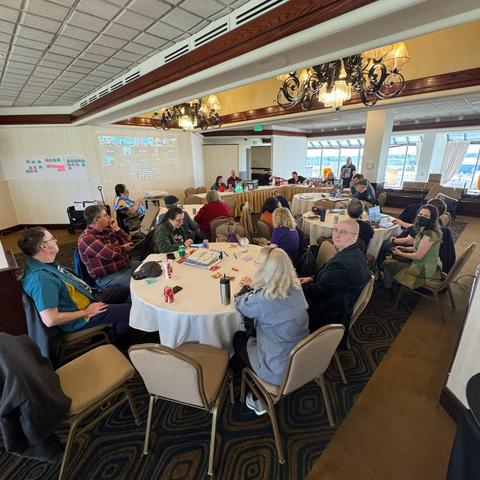 Norwescon’s staff gathered around tables in a hotel meeting room, with a Zoom window projects onto one wall.