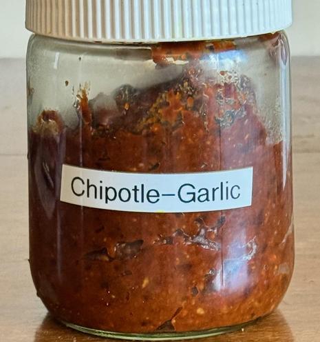 A jar with a homemade label that reads "Chipotle-garlic" and containing a red paste. Jar is squat and has a white cap.