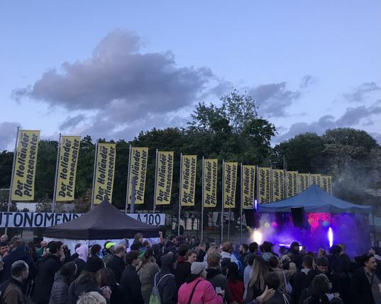 Eine Party auf der Straße, rechts ein pink leuchtender Pavillon. Im Hintergrund eine Reihe Fahnen des Pflanzenladens „Der Holländer“