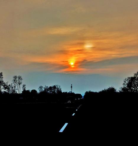 A low-angle shot shows a straight, dark road stretching into the distance towards a vibrant sunset. The sun, a bright orange orb, is partially obscured by clouds, casting a warm glow across the sky, which transitions from orange and yellow near the horizon to a muted grey-blue above. Silhouetted trees line both sides of the road, creating a dark border. White dashed lines mark the centre of the road, leading the eye towards the light. Two faint sets of headlights suggest vehicles in the distance.