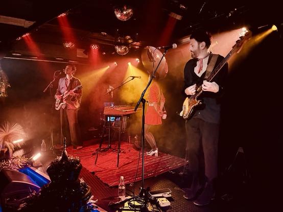 A live music performance featuring three musicians on stage. One guitarist sings while another plays bass. A keyboard is in the foreground, with ambient lighting and smoke effects creating a vibrant atmosphere. The performers are dressed in diverse styles, and a disco ball is visible