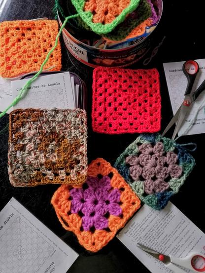Varios “cuadraditos de abuela” tejidos a crochet en colores vibrantes como naranja, fucsia, morado y verde, dispuestos sobre una mesa negra. Se ven tijeras, hojas impresas con instrucciones para tejer, y una lata reutilizada llena de lanas.