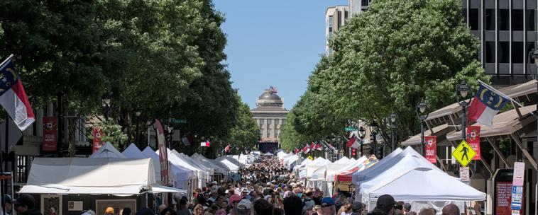 A photo of Fayetteville street looking towards the NC state capitol, during a festival. The street is packed with people, lined with white tents for various vendors.