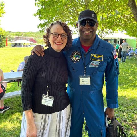 A photo of Mary Robinette Kowal and Leland Melvin. Mary Robinette is wearing a black turtleneck sweater and white, tan, and black striped linen pants, while Leland Melvin is wearing a blue astronaut jumpsuit covered with patches. They’re in the shade, and in the background is a picnic table plus multiple tents.