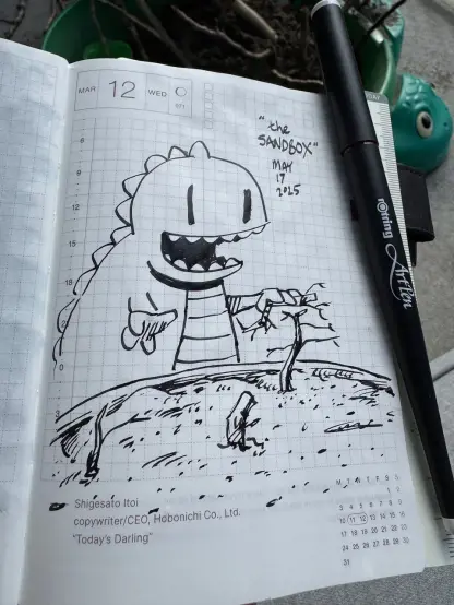 A photo of a planner with an ink line drawing cartoon of a Zilla playing with sticks in a sandbox.