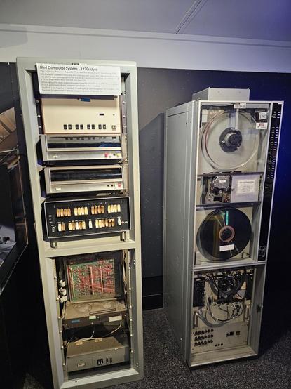 A photo of a vintage 1970s-style mini computer system on display in a museum. The setup includes two tall metal cabinets. The left cabinet houses various components such as switches, circuit boards, and a control panel with orange toggle switches and indicator lights. A sign at the top describes it as a "Mini Computer System – 1970s style," noting its use in atomic research. The right cabinet contains large reel-to-reel magnetic tape drives used for data storage, with visible cables and connectors at the bottom. Both units represent early computing technology.