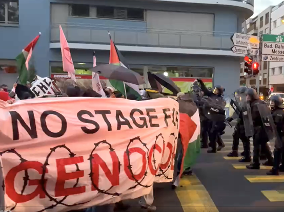 Screenshot van video Nau.ch: Demonstranten met heel groot spandoek waarop “NO STAGE FOR GENOCIDE”. In schemerlicht tegenover linie oproerpolitie.