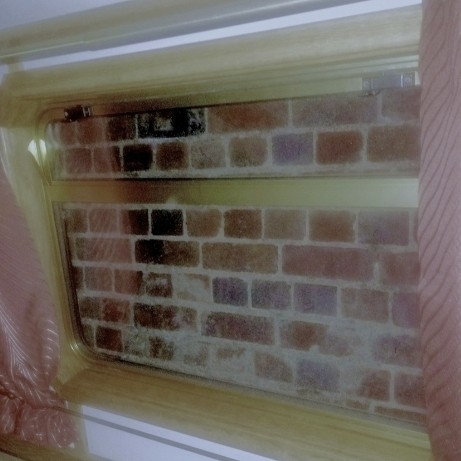 The view outside the boat window is … a solid, brick wall. The wall is only a few inches (10 centimeters) outside the window.

Inside, the window frame is brassy. The wall is painted white and the curtains are a dull red tint with faint, thin stripes.