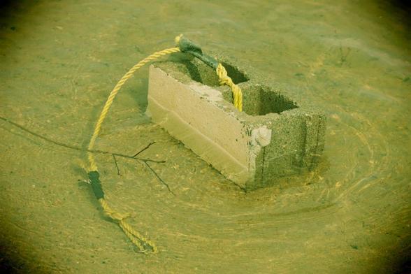 A concrete block rests in low clear water, attached to it is a short yellow, intended for looping onto a floating device. Next to the submerged portion of the rope is a very thin, short tree branch. The yellowy tinged green tone and vignetting emulates a plastic camera (in camera effect).