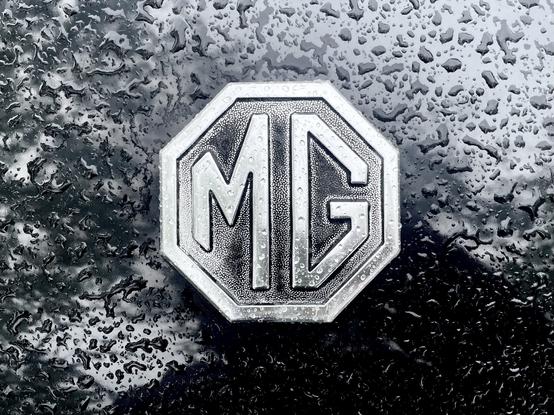 Photograph of an MG car logo consisting of a capital MG in an octagon. The logo is on a black car and covered by raindrops.