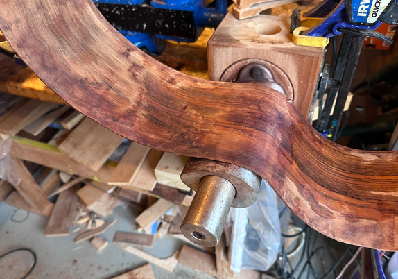 a messy workbench with some bent wood that will ultimately become the sides of a guitar. The wood is sitting on a bending iron - a heated tube of metal.