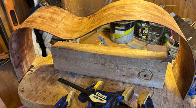 a messy workbench with some bent wood that will ultimately become the sides of a guitar - the side piece is close to the final shape.