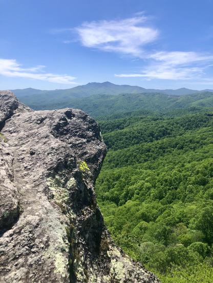 large rock outcrop on left overlooking a green forested valley below with blue peaks in the distance
