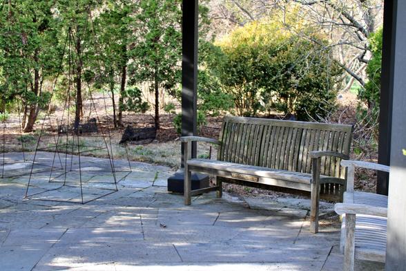 Benches on a patio under a shady pergola with trees and bushes in the background. There are metal four-sided frames as garden art next to the benches.