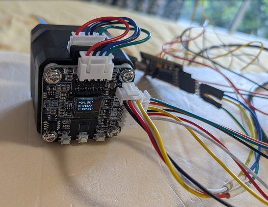 A photo of a stepper motor with various colourful wires attached. A small OLED screen on the back of the motor reads "-34.8k° 0.04err <-99Kclk".

Some trees are visible through a window in the background.