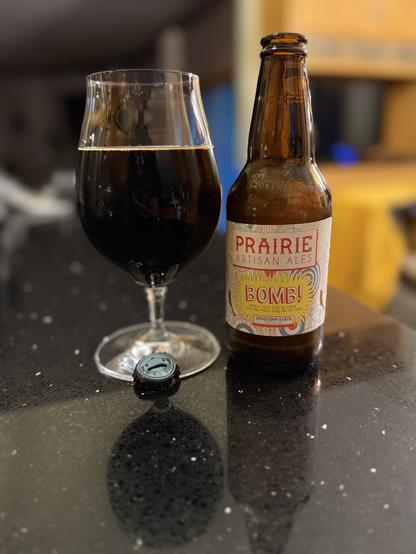 A tall snifter glass of dark beer is next to a brown bottle with a white label and lettering in light shades of red, blue, and yellow. The words “BOMB!” Is front and center.