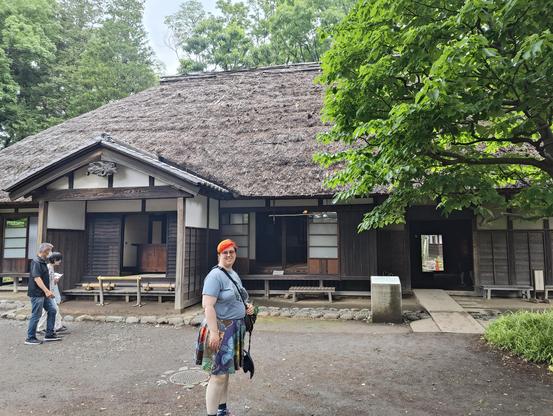 A photo of a person with bright rainbow hair and skirt standing in front of an antique thatched roof building